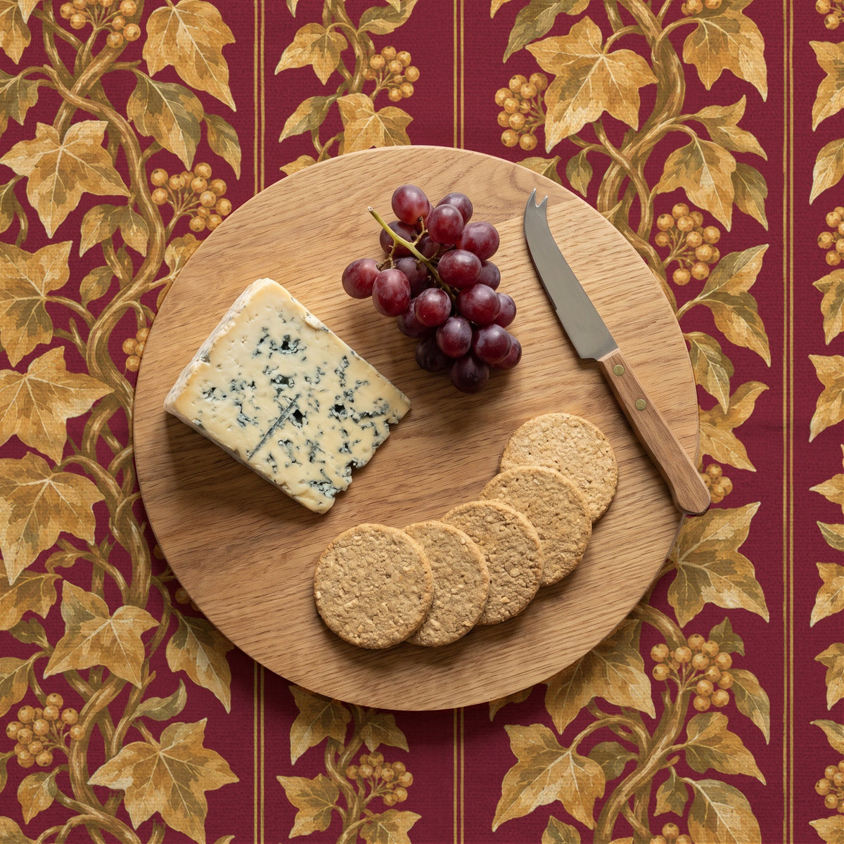 Burgundy and gold Gilded Ivy Christmas tablecloth with an oak cheeseboard, blue cheese, grapes and oatcakes