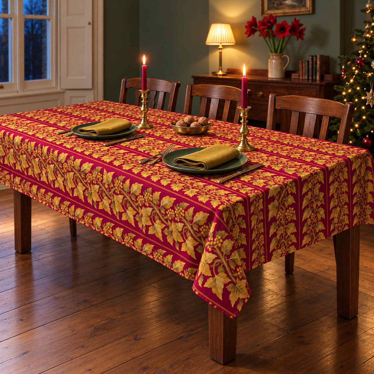 Burgundy and gold Gilded Ivy Christmas tablecloth with a soft drape on a rectangular wooden table with visible legs, green plates, gold napkins and candlelight.