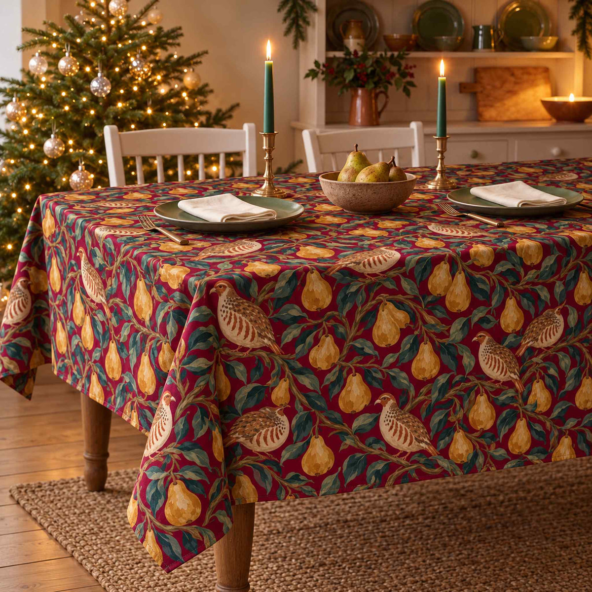 Burgundy Partridge and Pear Christmas tablecloth draped over a dining table with two ivory place settings, folded napkins and a small bowl of pears.