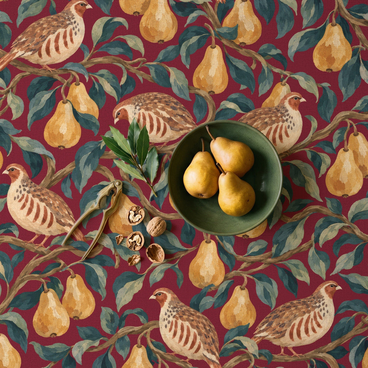 Partridge and Pear Christmas tablecloth in burgundy seen from above, with a green stoneware bowl of three pears, a brass nutcracker, walnuts and a sprig of bay