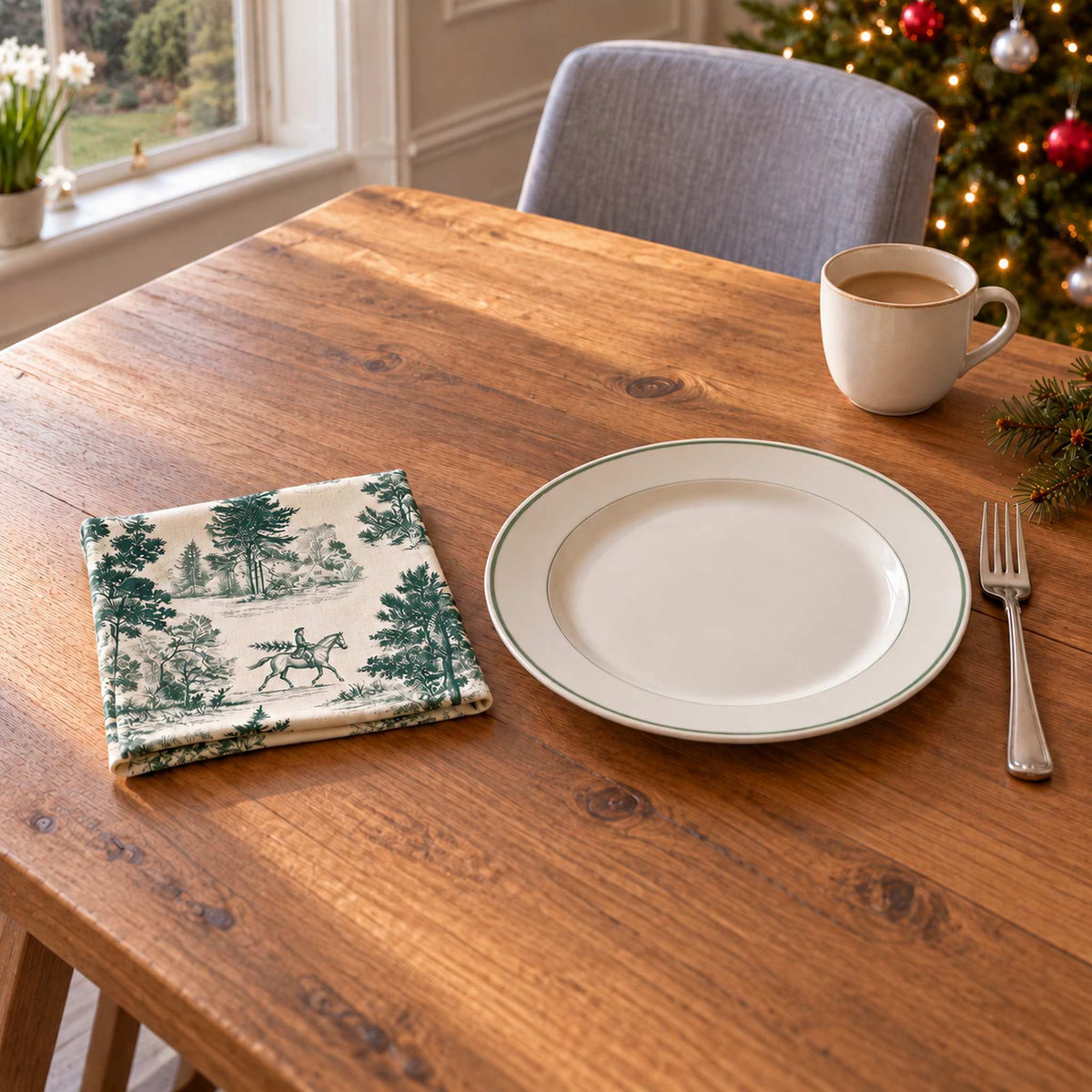 Green on ivory Fir Forest Toile cotton napkin in a compact square fold beside a dinner plate and coffee cup on a wooden table in daylight.