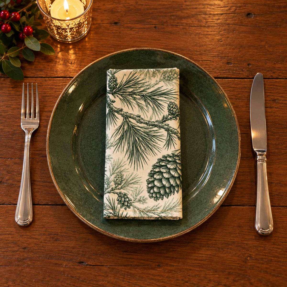 Green on ivory Pine Bough cotton napkin neatly folded on a dinner plate with cutlery and candlelight on a wooden Christmas table.