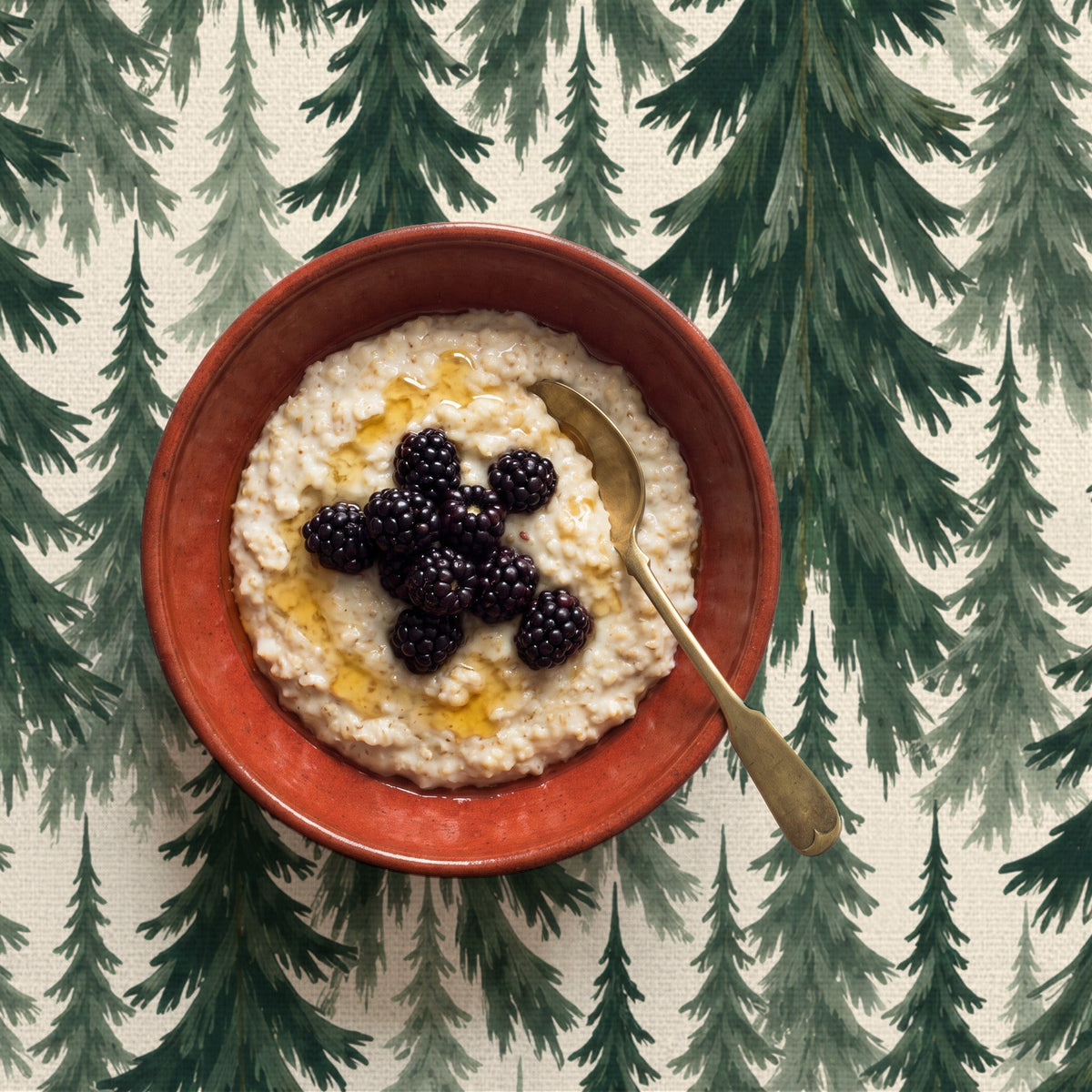 Spruce Stand Christmas tablecloth in green on ivory seen from above, with a rust-red bowl of porridge topped with blackberries and honey and a brass spoon