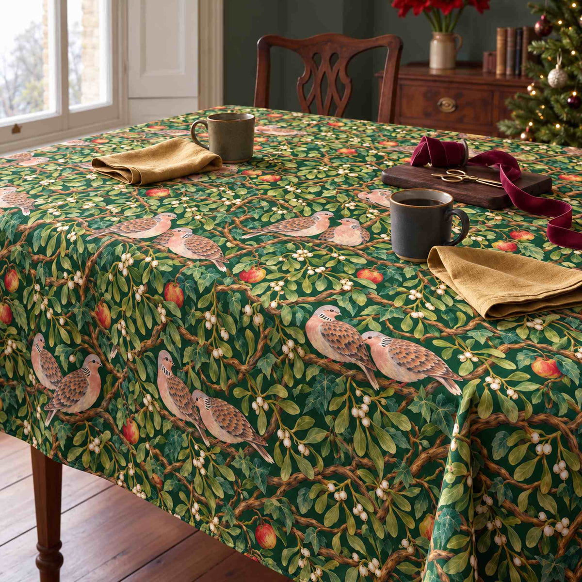 Green Turtle Doves and Mistletoe Christmas tablecloth draped over a dining table with mugs, gold napkins and wrapping ribbon in soft winter daylight.