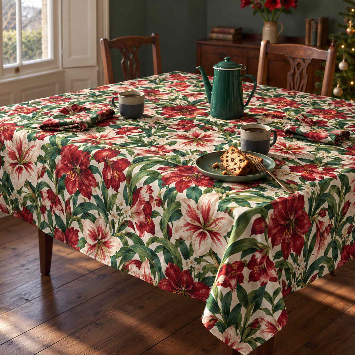 Ivory Amaryllis Christmas tablecloth draped over a dining table with a green coffee pot, mugs and matching floral napkins in soft winter daylight.