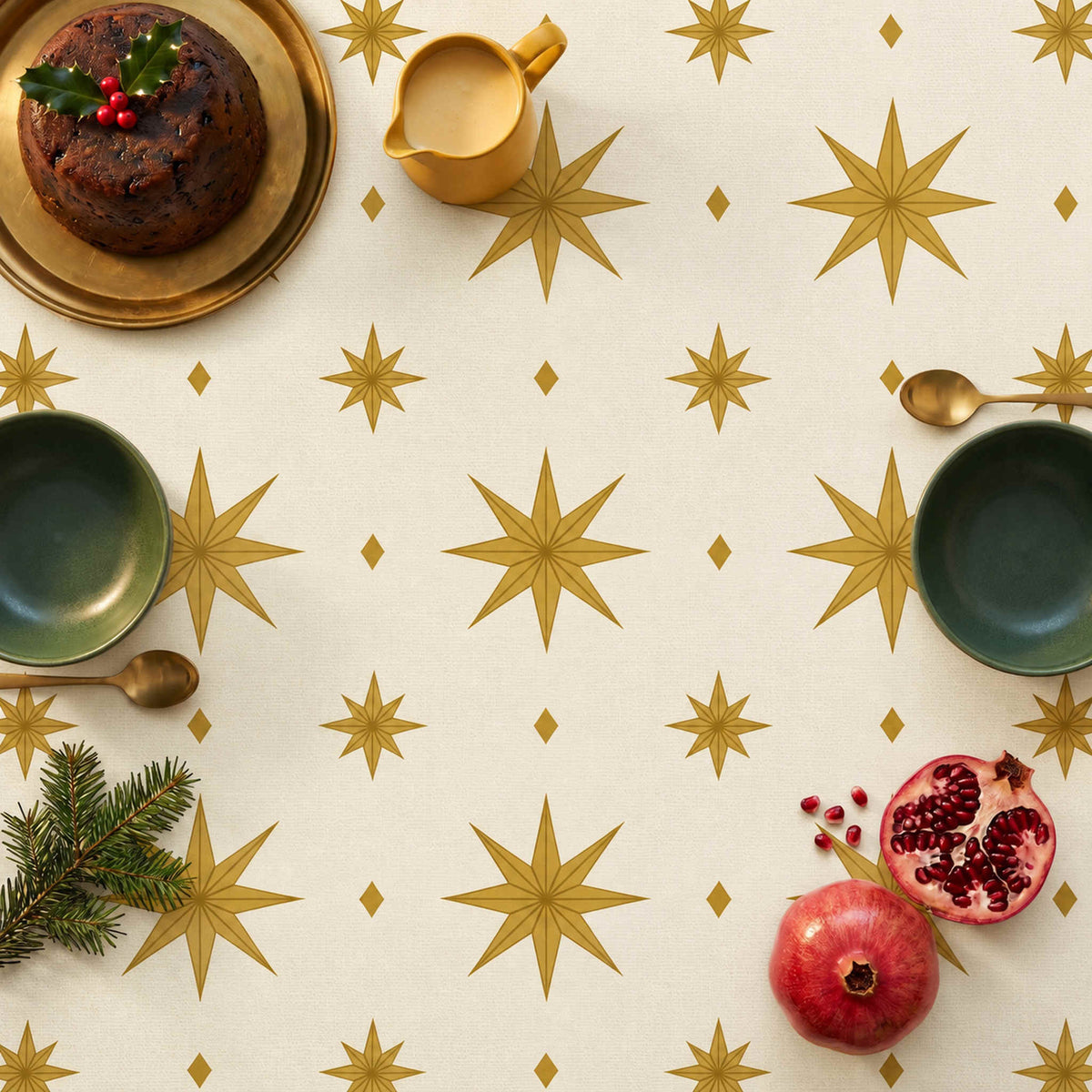 Ivory and gold Stars Christmas tablecloth seen from above, styled with Christmas pudding on a brass platter, a mustard custard jug, green bowls, brass spoons, pomegranates and fir.