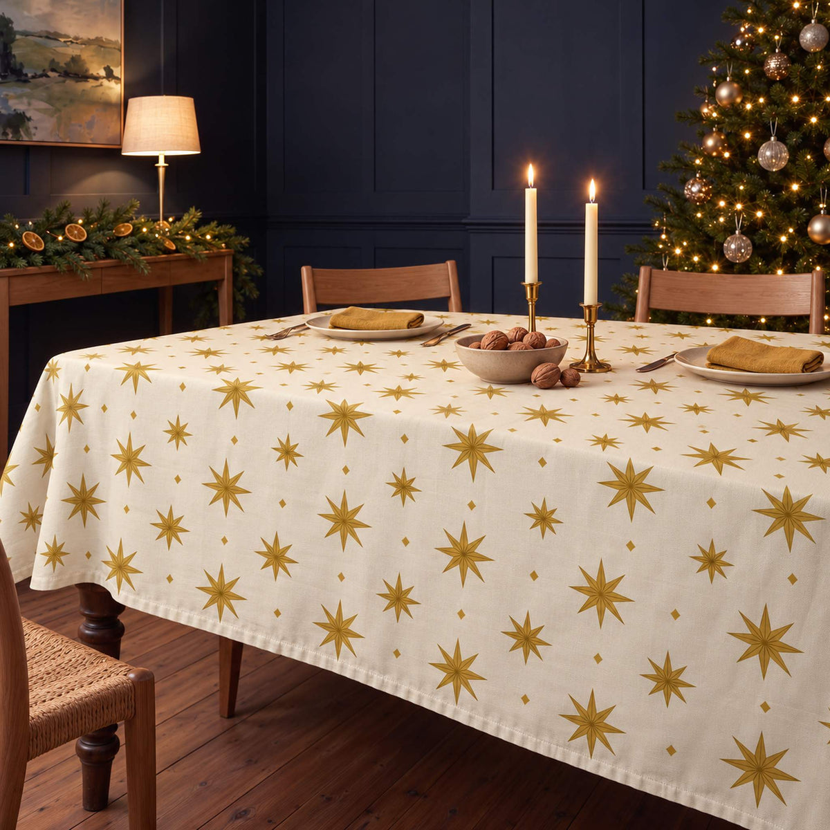 Ivory and gold Stars Christmas tablecloth draped over a dining table with gold napkins, ivory plates, walnuts and candles in a navy-panelled room.