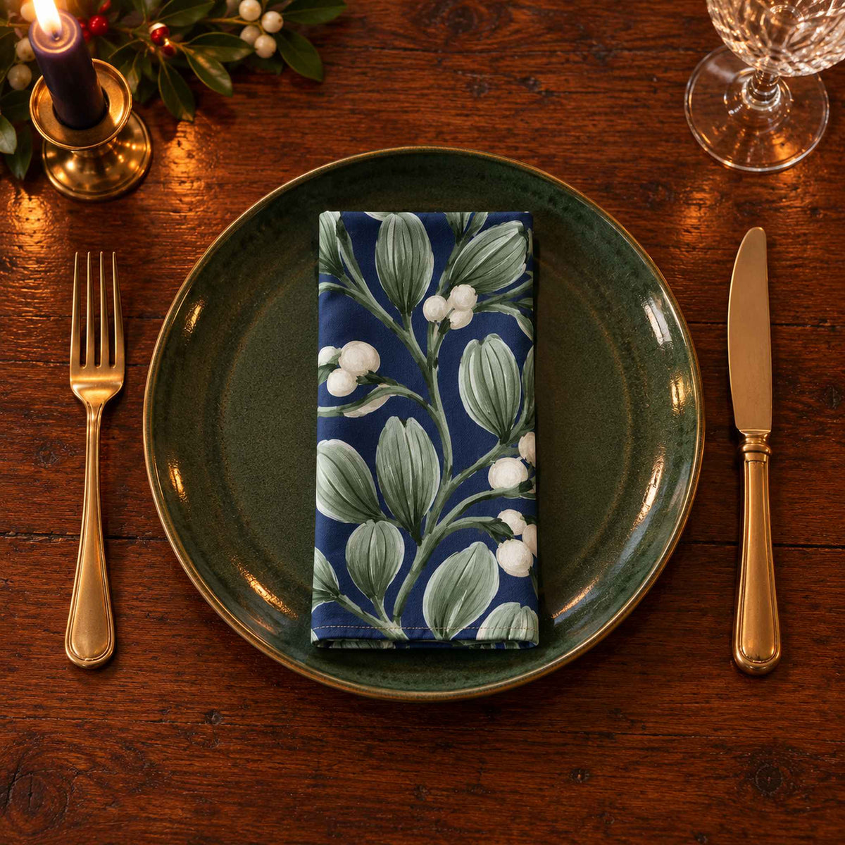 Navy Mistletoe cotton napkin neatly folded on a dinner plate with cutlery and candlelight on a wooden Christmas table.