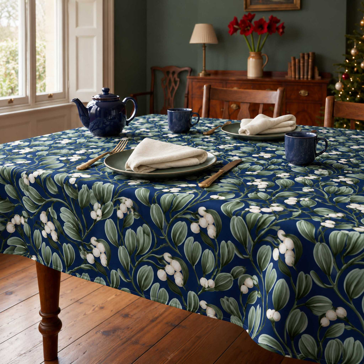 Navy Mistletoe Christmas tablecloth draped over a dining table with a navy teapot, two mugs and ivory napkins in soft winter daylight.