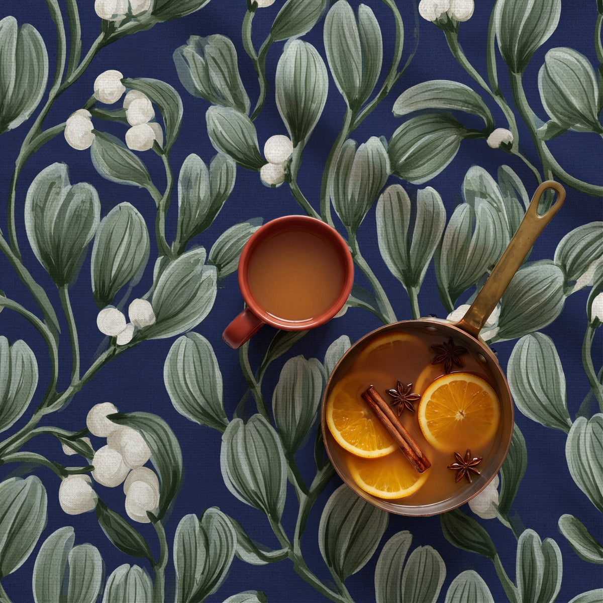 Navy Mistletoe Christmas tablecloth with sage green leaves and white berries, seen from above with a copper pan of mulled cider and a rust-red mug.
