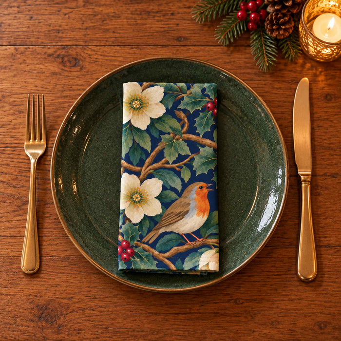 Navy Robins and Christmas Roses cotton napkin neatly folded on a dinner plate with cutlery and candlelight on a wooden Christmas table.