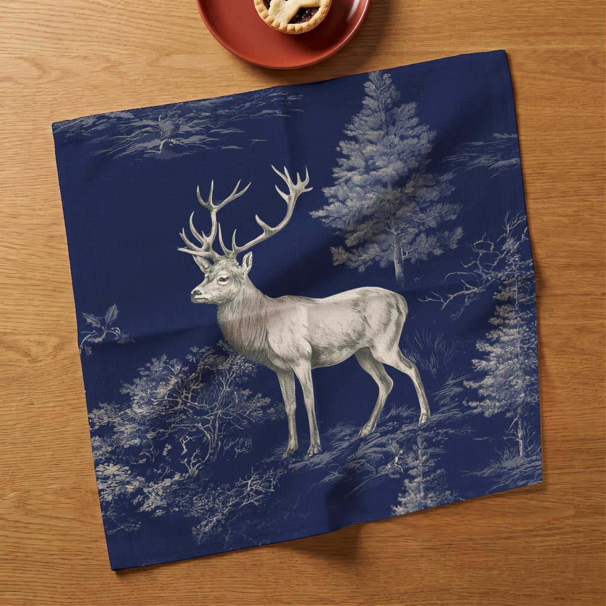 Navy Stag Woodland Christmas napkin opened face up on an oak table, showing a complete stag and woodland print beside a mince pie on a red plate