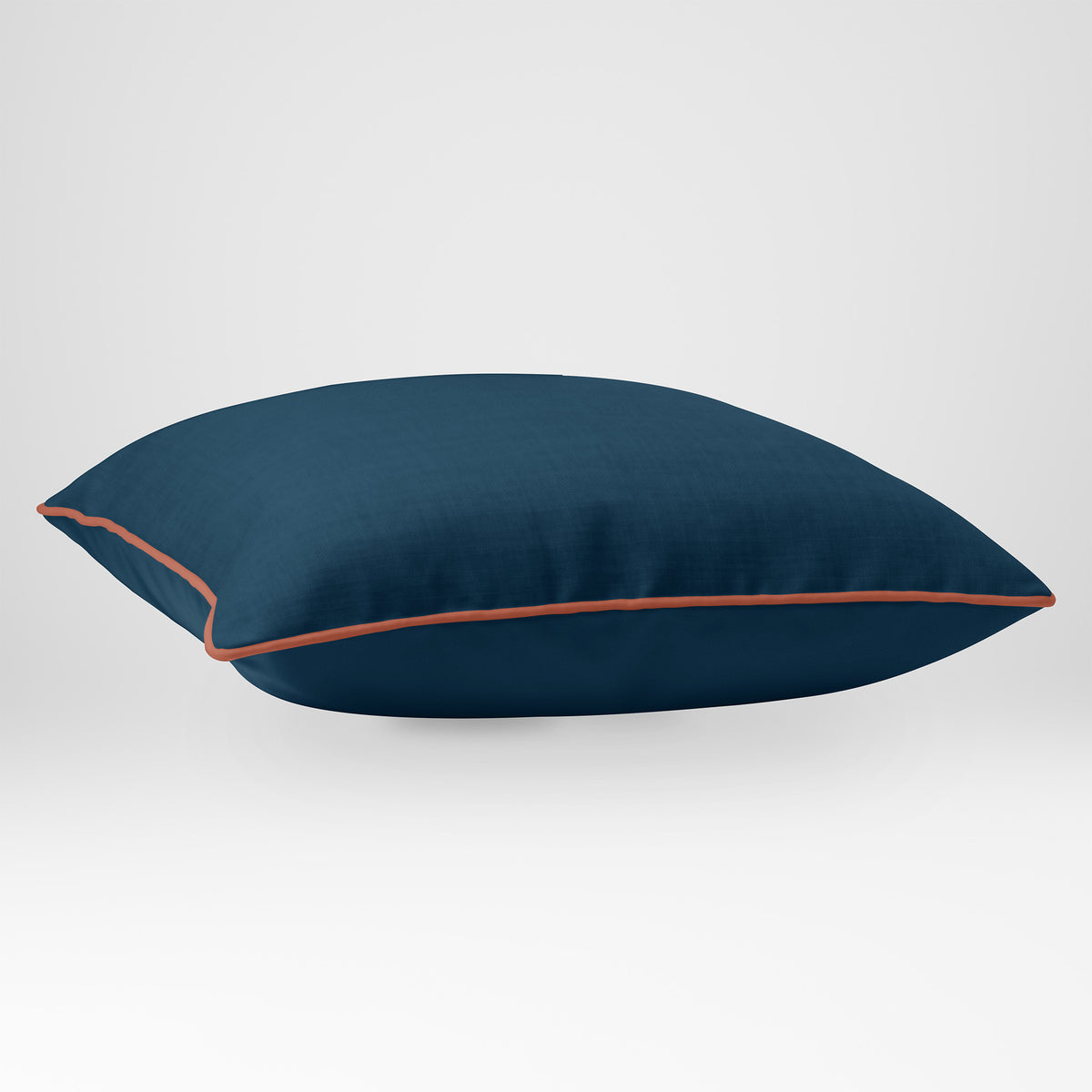 Angled view showing the front and Deep Teal plain reverse of an Avara plain square cushion in Deep Teal, finished with Persimmon piping.