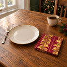 Burgundy and gold Gilded Ivy cotton napkin in a compact square fold beside a dinner plate and coffee cup on a wooden table in daylight.