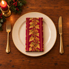 Burgundy and gold Gilded Ivy cotton napkin neatly folded on a dinner plate with cutlery and candlelight on a wooden Christmas table.