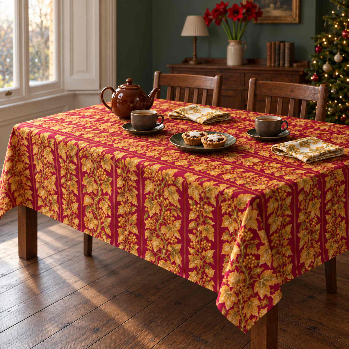 Burgundy and gold Gilded Ivy Christmas tablecloth on a rectangular wooden dining table, styled with a brown teapot, two cups and mince pies in a green room.