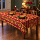 Burgundy and gold Gilded Ivy Christmas tablecloth with a soft drape on a rectangular wooden table with visible legs, green plates, gold napkins and candlelight.