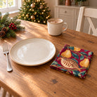 Burgundy Partridge and Pear cotton napkin in a compact square fold beside a dinner plate and coffee cup on a wooden table in daylight.