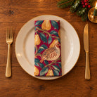 Burgundy Partridge and Pear cotton napkin neatly folded on a dinner plate with cutlery and candlelight on a wooden Christmas table.
