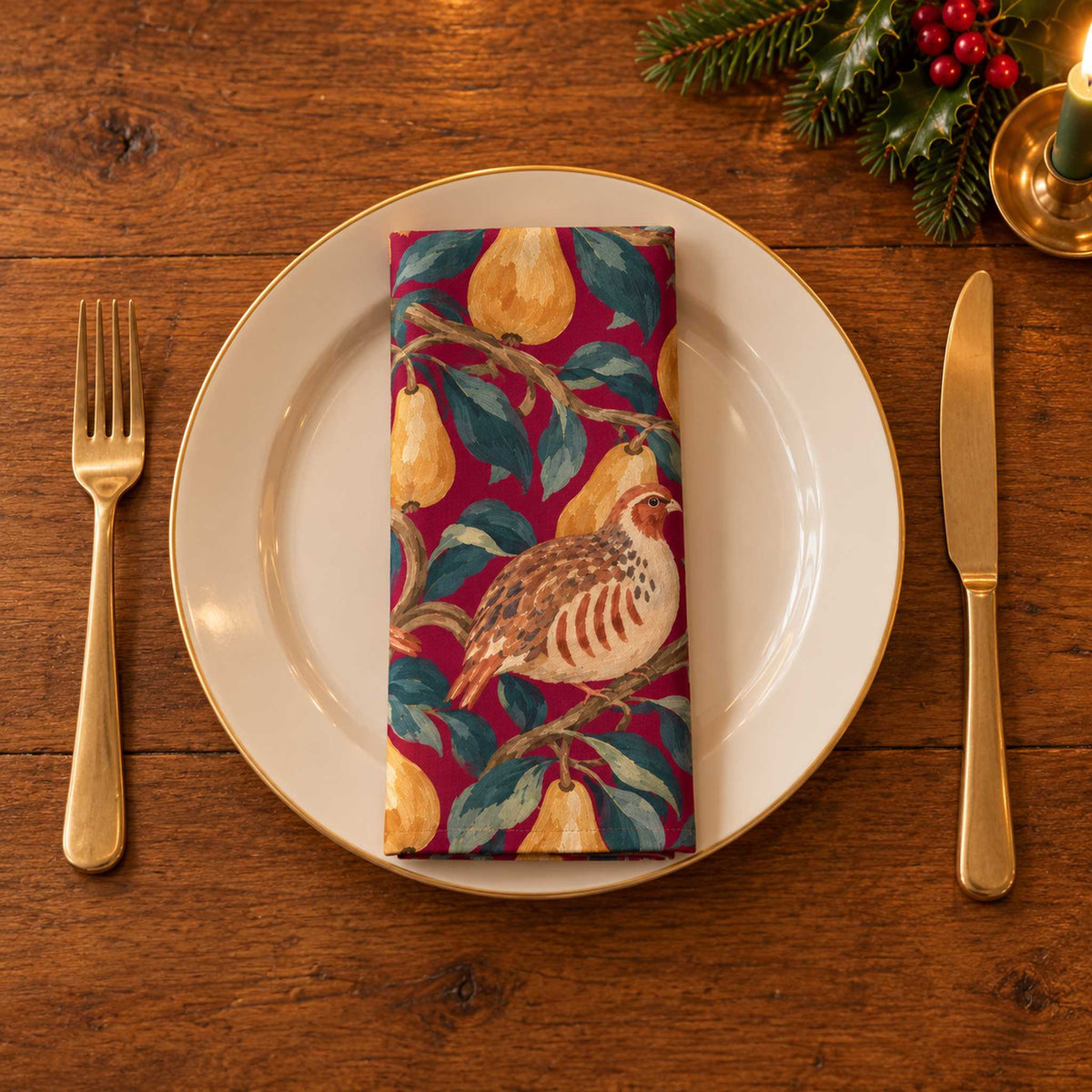 Burgundy Partridge and Pear cotton napkin neatly folded on a dinner plate with cutlery and candlelight on a wooden Christmas table.