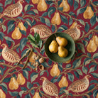 Partridge and Pear Christmas tablecloth in burgundy seen from above, with a green stoneware bowl of three pears, a brass nutcracker, walnuts and a sprig of bay