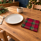 Burgundy Tartan cotton napkin in a compact square fold beside a dinner plate and coffee cup on a wooden table in daylight.