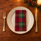 Burgundy Tartan cotton napkin neatly folded on a dinner plate with cutlery and candlelight on a wooden Christmas table.