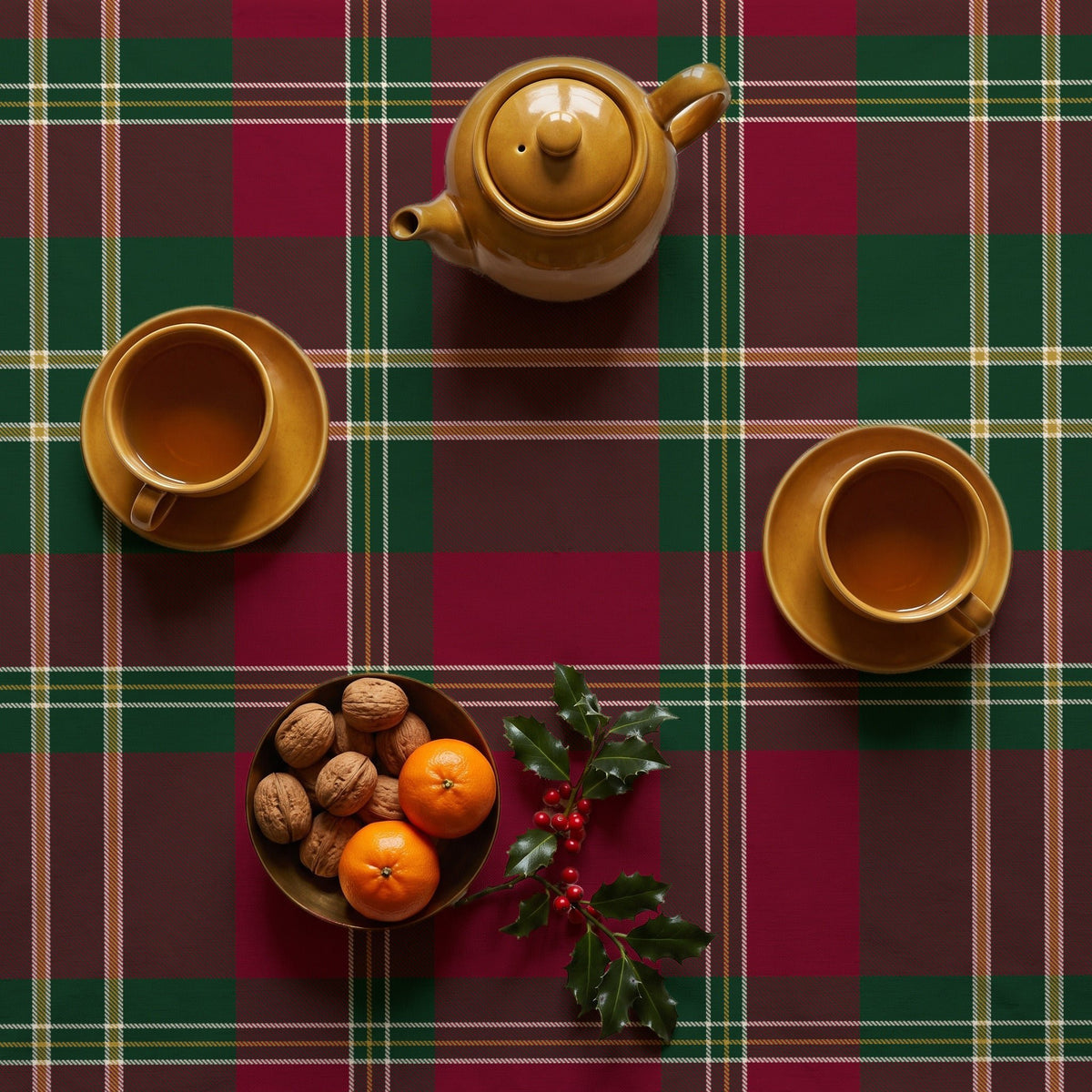Burgundy tartan Christmas tablecloth seen from above, set for afternoon tea with a mustard teapot, two cups of tea, a bowl of walnuts and clementines and a sprig of holly