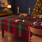 Burgundy Tartan Christmas tablecloth draped over a dining table with matching tartan napkins, ivory plates, evergreen branches and lit candles.