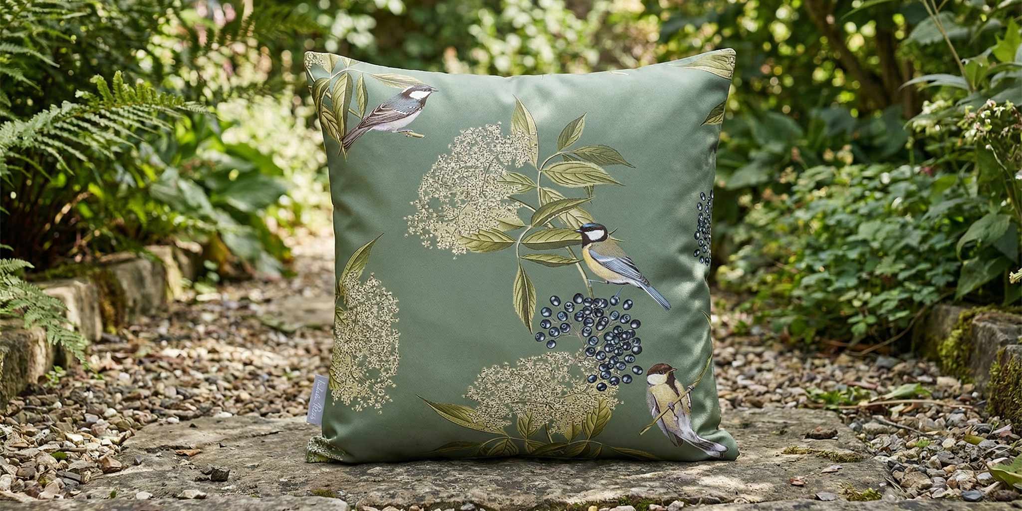 Green outdoor cushion with bird and elderflower print, styled on a stone path in a garden setting.