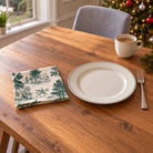 Green on ivory Fir Forest Toile cotton napkin in a compact square fold beside a dinner plate and coffee cup on a wooden table in daylight.