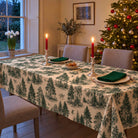 Green on ivory Fir Forest Toile Christmas tablecloth draped over a dining table with ivory plates, green napkins and red candles beside a Christmas tree.