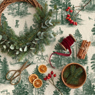 Fir Forest Toile Christmas tablecloth in green on ivory seen from above, with a half-made wreath of fir and eucalyptus, brass scissors, red velvet ribbon, cinnamon sticks, dried orange slices, rosehips and a wooden bowl of fir sprigs