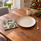 Green on ivory Pine Bough cotton napkin in a compact square fold beside a dinner plate and coffee cup on a wooden table in daylight.