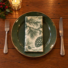Green on ivory Pine Bough cotton napkin neatly folded on a dinner plate with cutlery and candlelight on a wooden Christmas table.