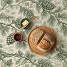 Pine Bough Christmas tablecloth in green on ivory seen from above, set for a slow winter breakfast with a sourdough loaf on an oak board, butter with a brass knife and a pot of marmalade