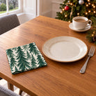 Green on ivory Spruce Stand cotton napkin in a compact square fold beside a dinner plate and coffee cup on a wooden table in daylight.