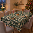 Spruce Stand Christmas tablecloth in green on ivory, shown from a raised three-quarter view over a rectangular dining table with grey chairs and red candles.