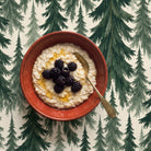Spruce Stand Christmas tablecloth in green on ivory seen from above, with a rust-red bowl of porridge topped with blackberries and honey and a brass spoon