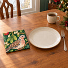 Green Turtle Doves and Mistletoe cotton napkin in a compact square fold beside a dinner plate and coffee cup on a wooden table in daylight.
