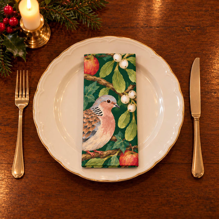 Green Turtle Doves and Mistletoe cotton napkin neatly folded on a dinner plate with cutlery and candlelight on a wooden Christmas table.