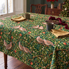 Green Turtle Doves and Mistletoe Christmas tablecloth draped over a dining table with mugs, gold napkins and wrapping ribbon in soft winter daylight.