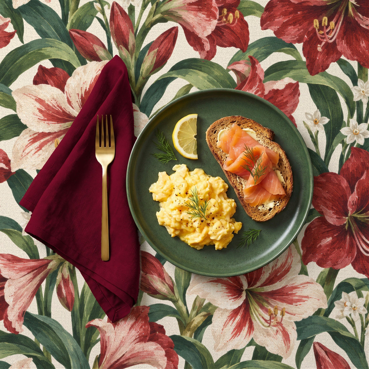 Amaryllis Christmas tablecloth in ivory seen from above, set for Christmas morning breakfast with a green plate of scrambled eggs and smoked salmon on brown toast, and a folded burgundy napkin with a brass fork