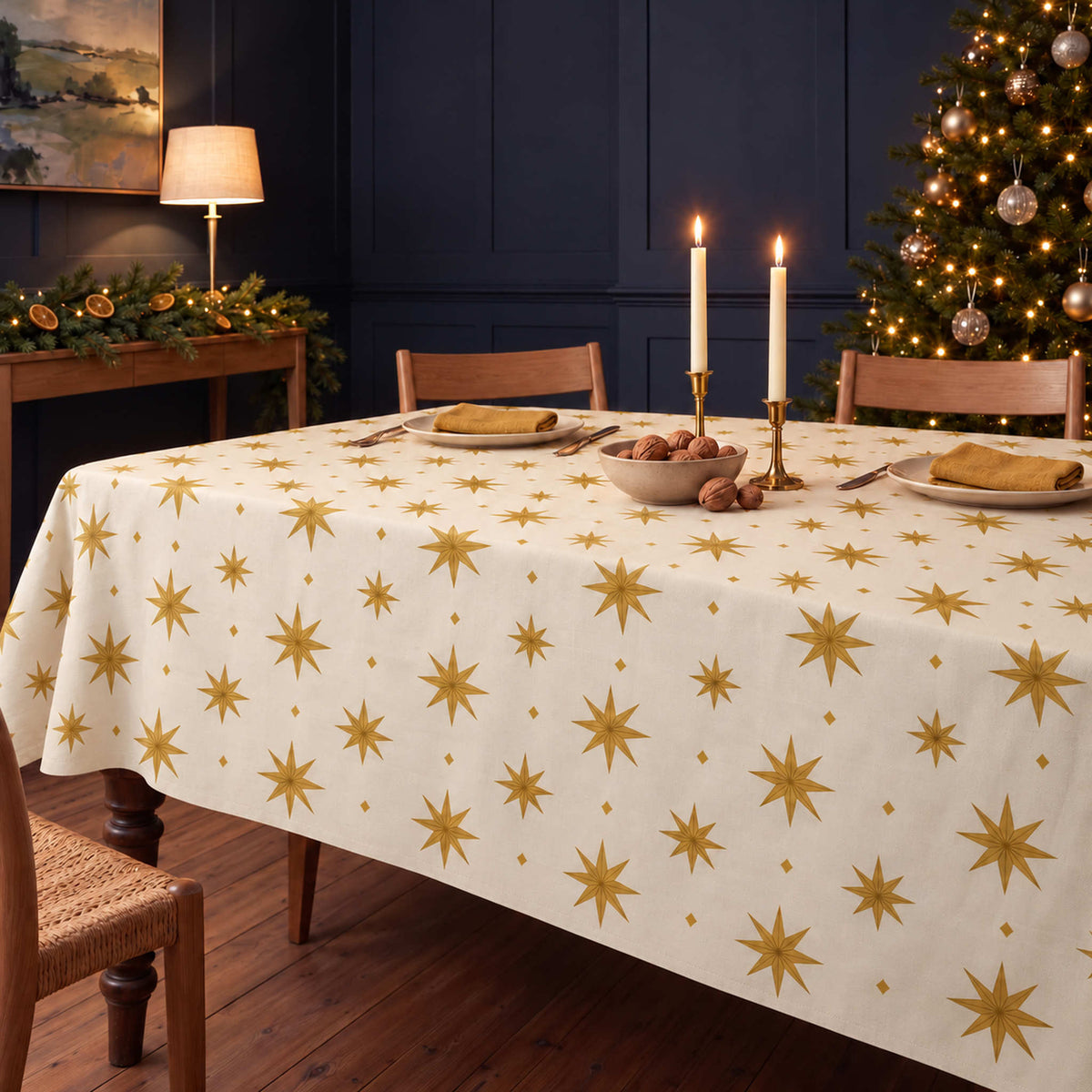 Ivory and gold Stars Christmas tablecloth draped over a dining table with gold napkins, ivory plates, walnuts and candles in a navy-panelled room.