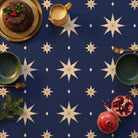 Stars Christmas tablecloth in navy and gold seen from above, set for Christmas pudding with a holly-topped pudding on a brass platter, a mustard jug of custard, two green bowls with brass spoons, pomegranates and a sprig of fir