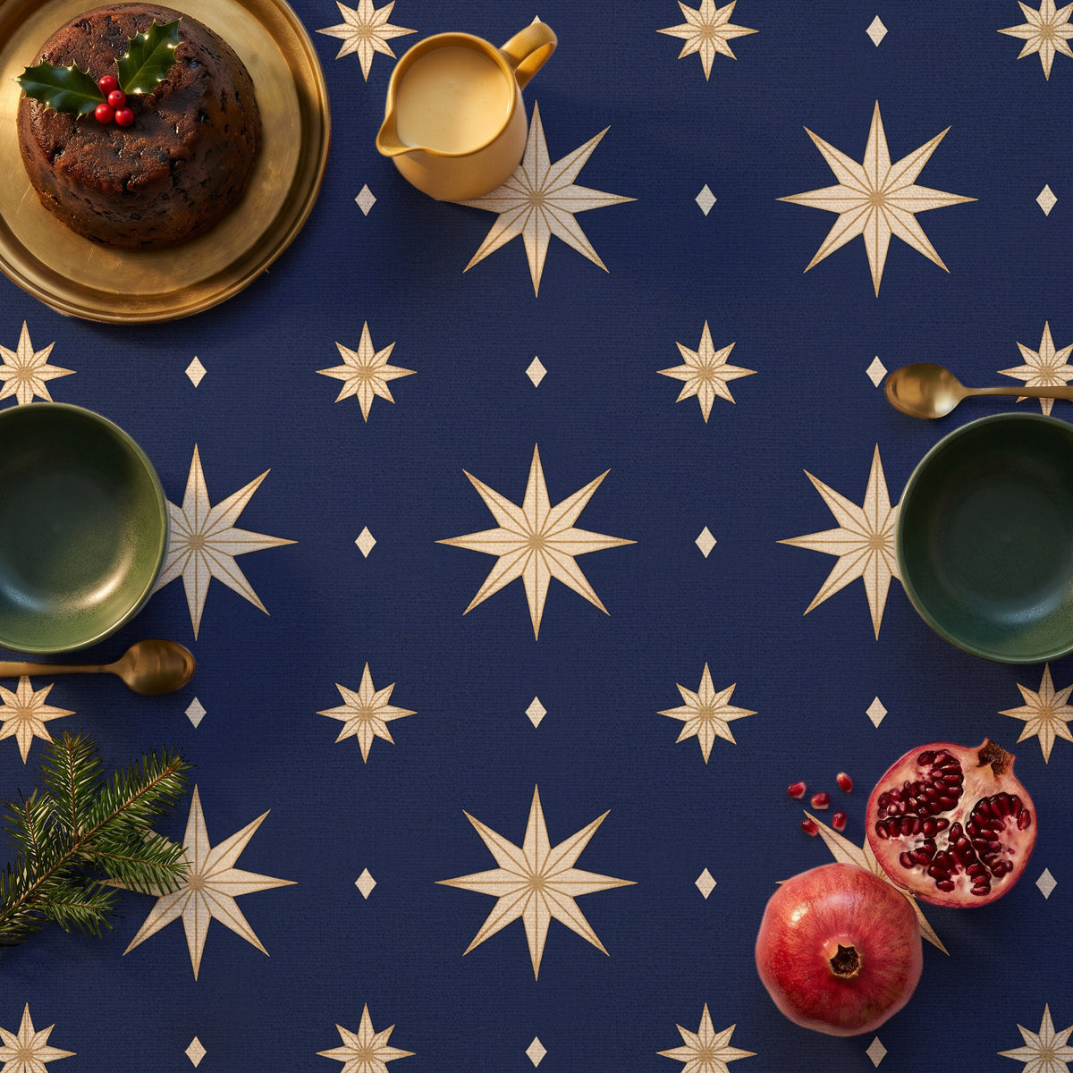 Stars Christmas tablecloth in navy and gold seen from above, set for Christmas pudding with a holly-topped pudding on a brass platter, a mustard jug of custard, two green bowls with brass spoons, pomegranates and a sprig of fir