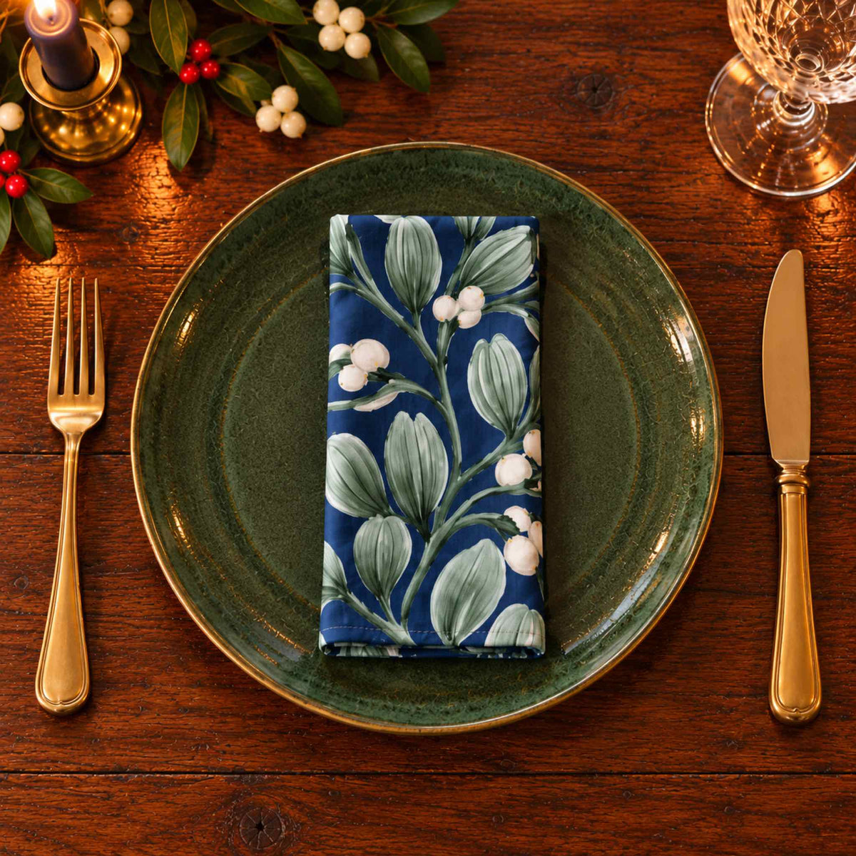 Close overhead view of a navy Mistletoe cotton Christmas napkin folded on a green dinner plate, with brass cutlery and candlelight.