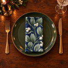 Navy Mistletoe cotton napkin neatly folded on a dinner plate with cutlery and candlelight on a wooden Christmas table.