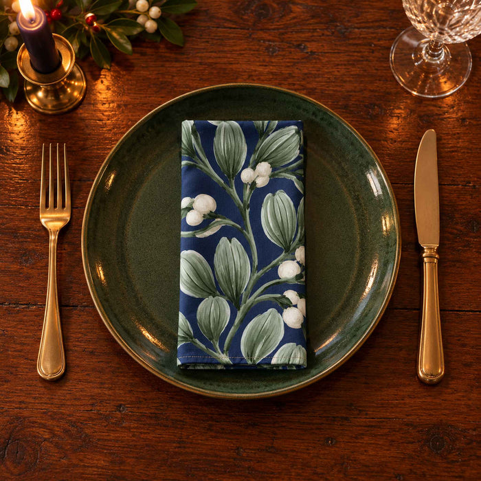 Navy Mistletoe cotton napkin neatly folded on a dinner plate with cutlery and candlelight on a wooden Christmas table.