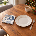 Navy on ivory Village Toile cotton napkin in a compact square fold beside a dinner plate and coffee cup on a wooden table in daylight.